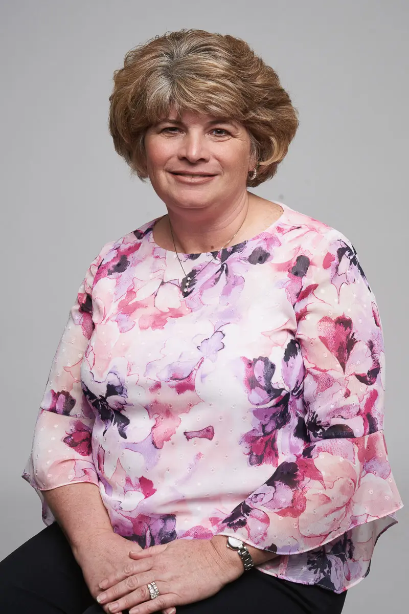 A woman with short, light brown hair wearing a pink, white, and purple floral blouse sits and smiles at the camera against a plain light gray background.