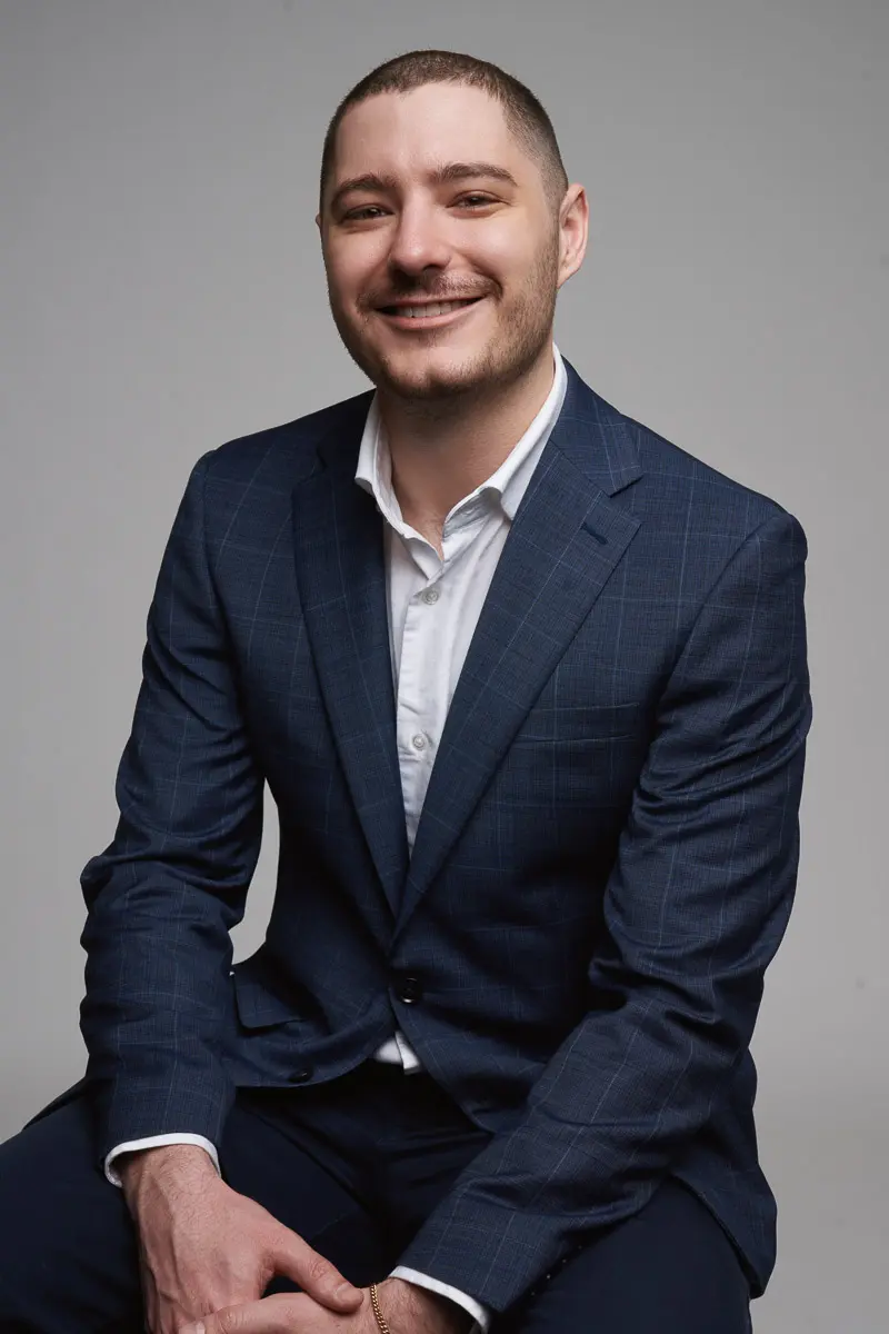 A man with short hair and a trimmed beard, wearing a dark blue suit and white shirt, sits and smiles at the camera against a plain gray background.
