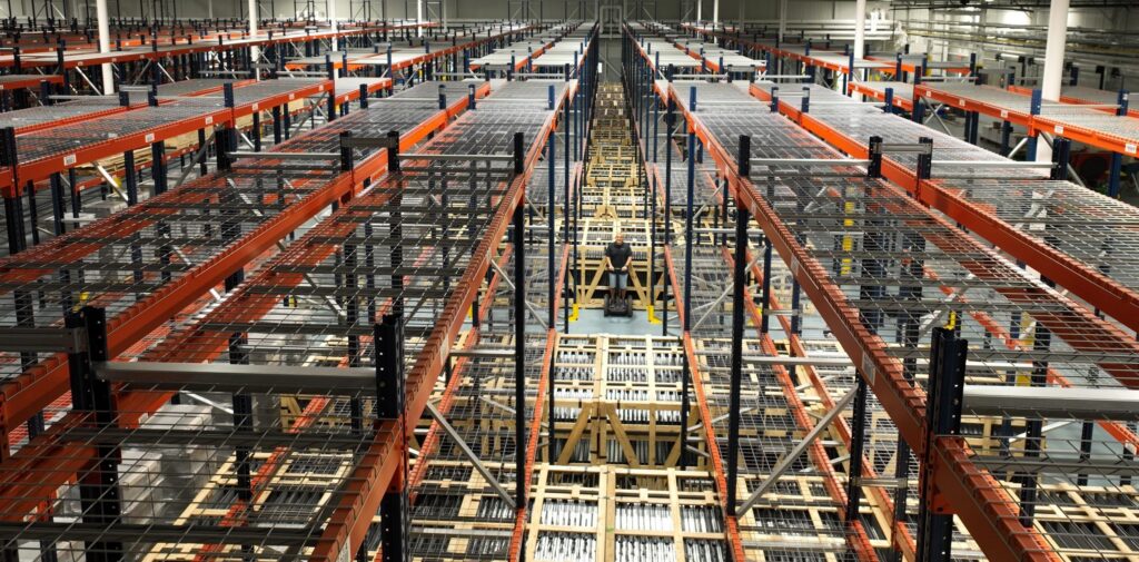 Bird’s-eye view of an empty warehouse featuring long rows of metal storage racks and shelves, with bright lighting and an organized, industrial atmosphere. No products or items are stored on the racks.