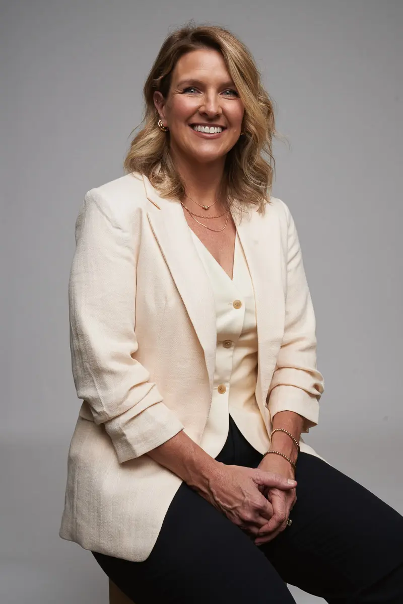 A woman with shoulder-length blonde hair, wearing a cream-colored blazer and top with dark pants, sits on a stool and smiles at the camera against a plain background.