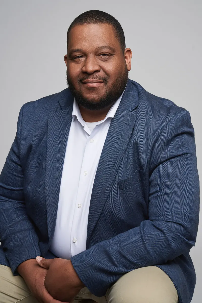 A man with short hair and a beard, wearing a blue blazer, white shirt, and khaki pants, sits and smiles slightly against a plain gray background.