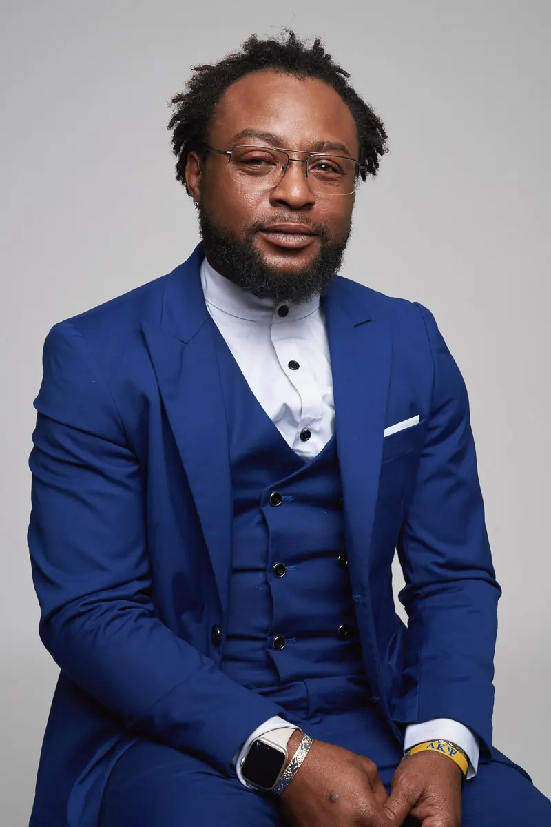 A man with a beard and glasses wears a royal blue suit, white dress shirt, and wristwatch. He is seated against a plain light gray background, looking at the camera with a slight smile.