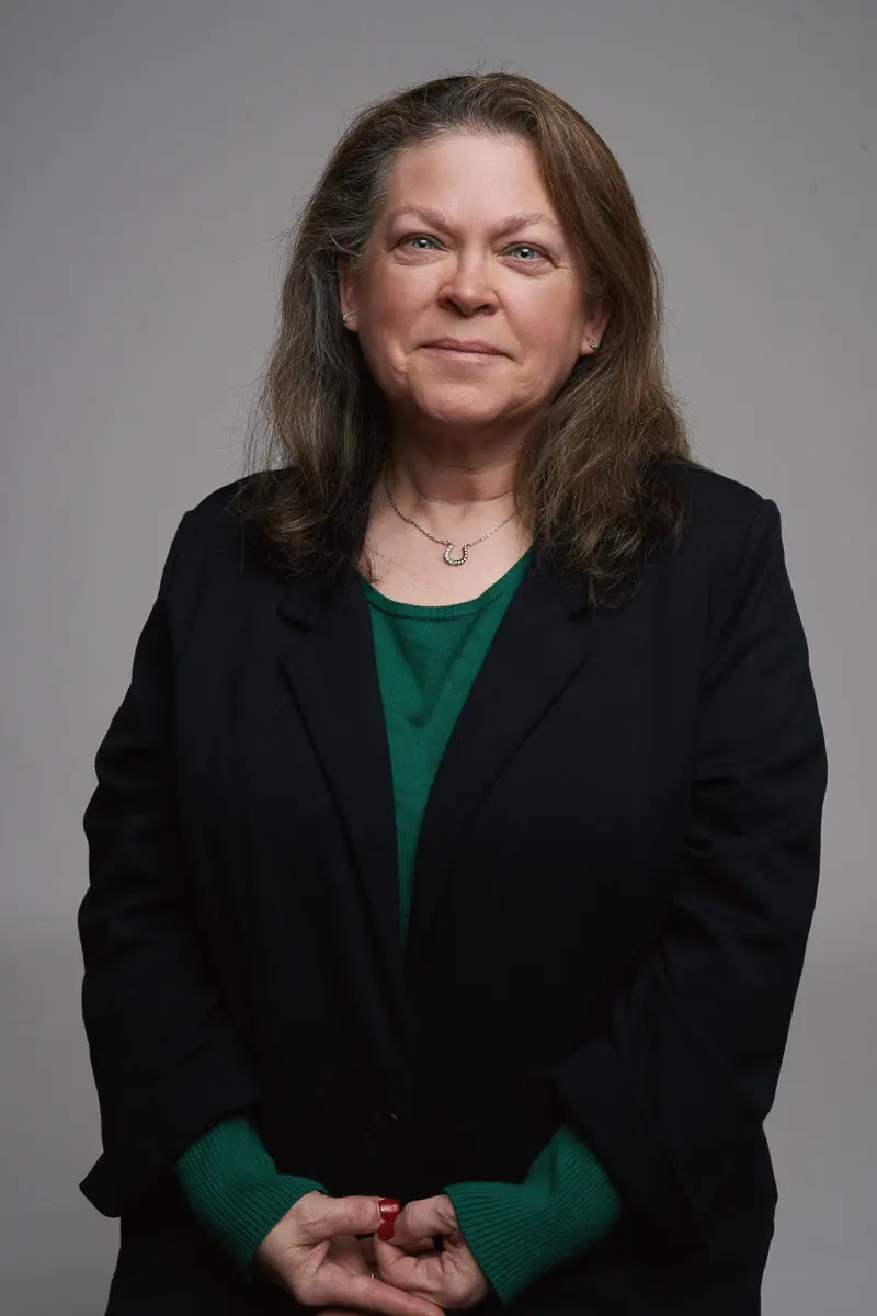 A woman with long brown hair wearing a dark blazer over a green top stands against a plain gray background, smiling softly with her hands clasped in front of her.