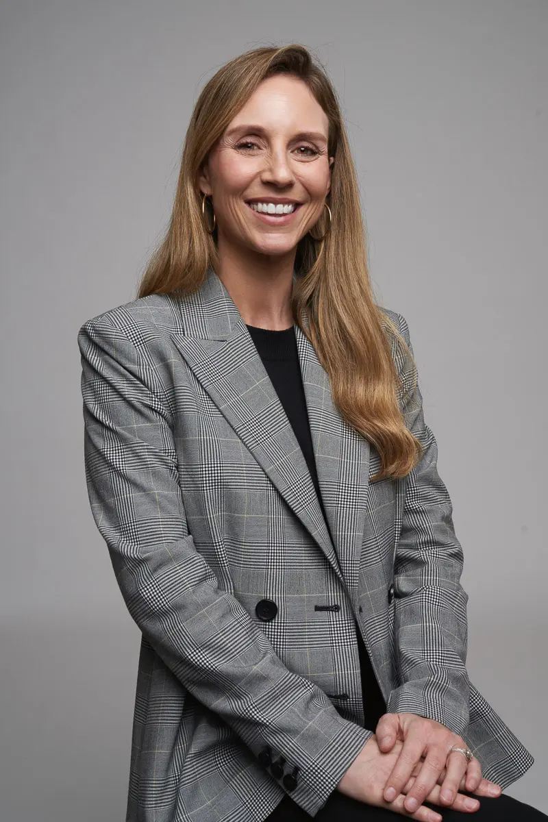 A woman with long blonde hair smiles, standing with her hands folded. She is wearing a grey plaid blazer over a black top against a plain, light grey background.
