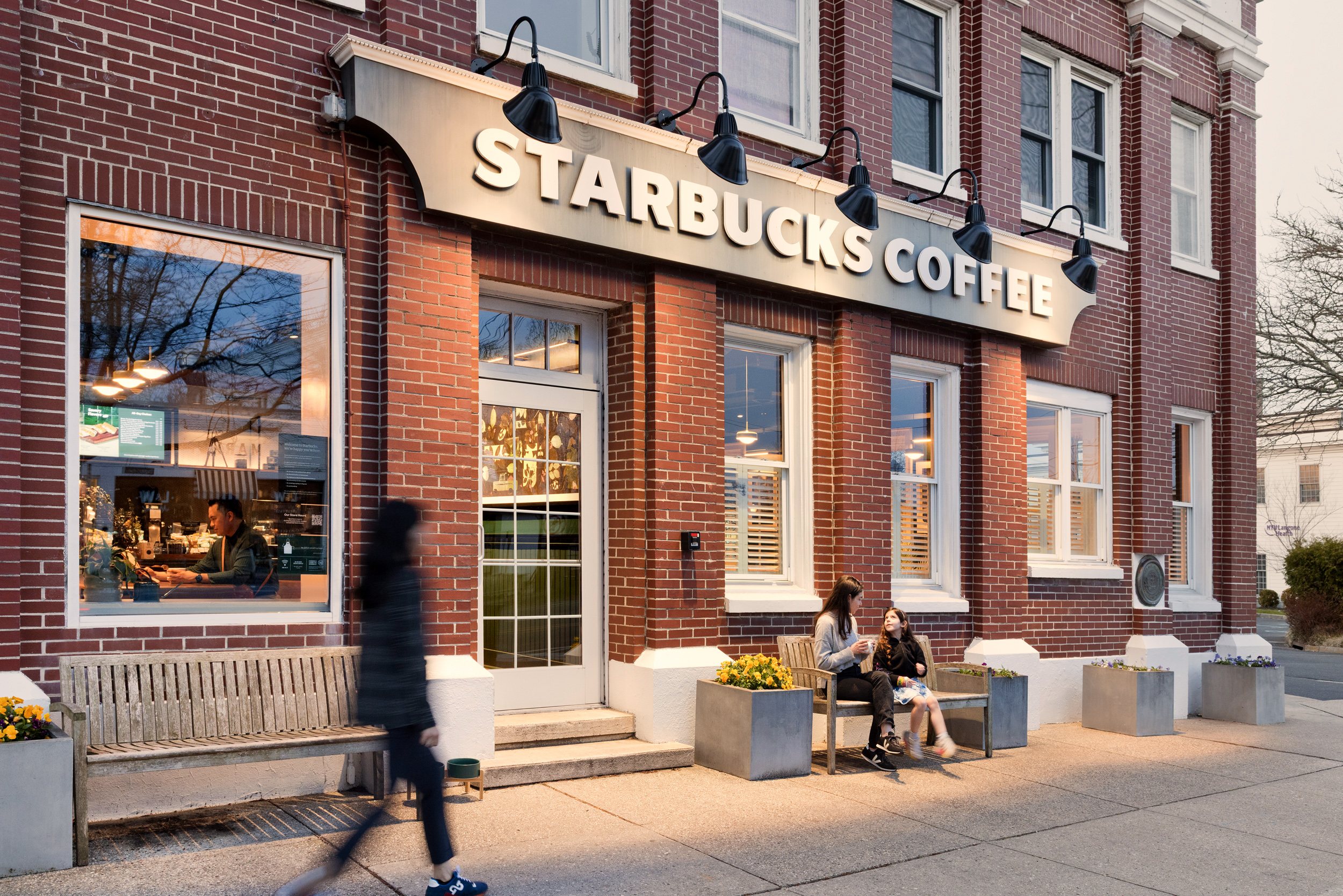 A brick building with a large "Starbucks Coffee" sign above the entrance. Two people sit outside on a bench, drinking coffee, while another person walks by. The shop’s interior is visible through the windows.