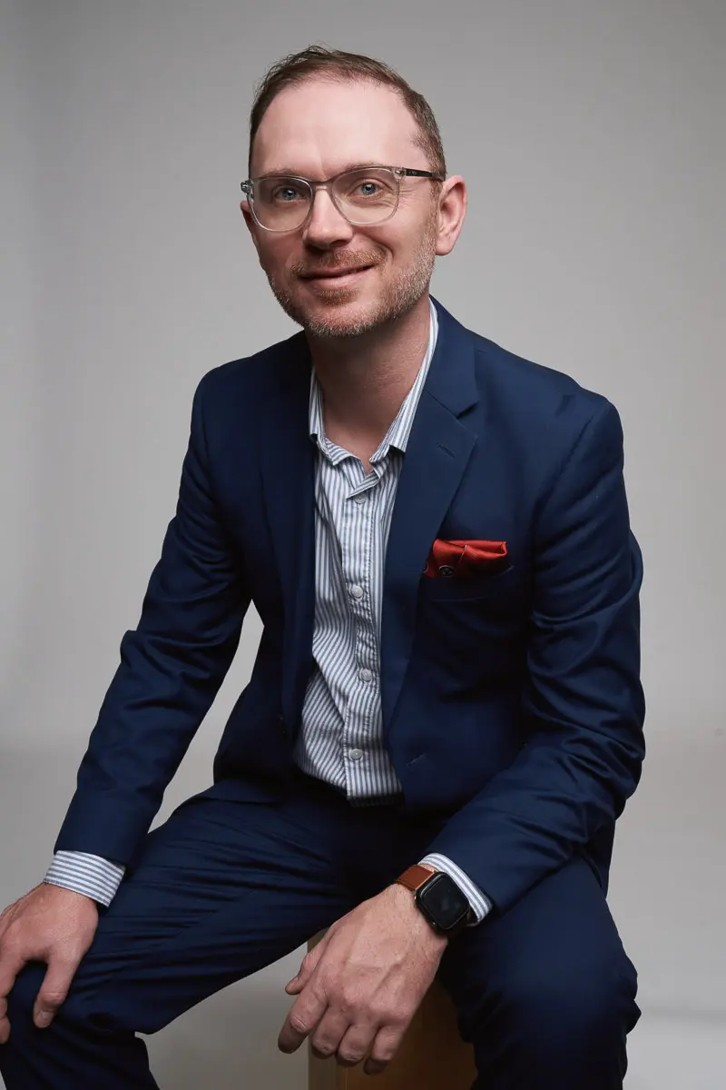 A man wearing glasses and a navy blue suit with a red pocket square sits on a stool against a plain background, smiling slightly at the camera. He has short hair and a smartwatch on his wrist.