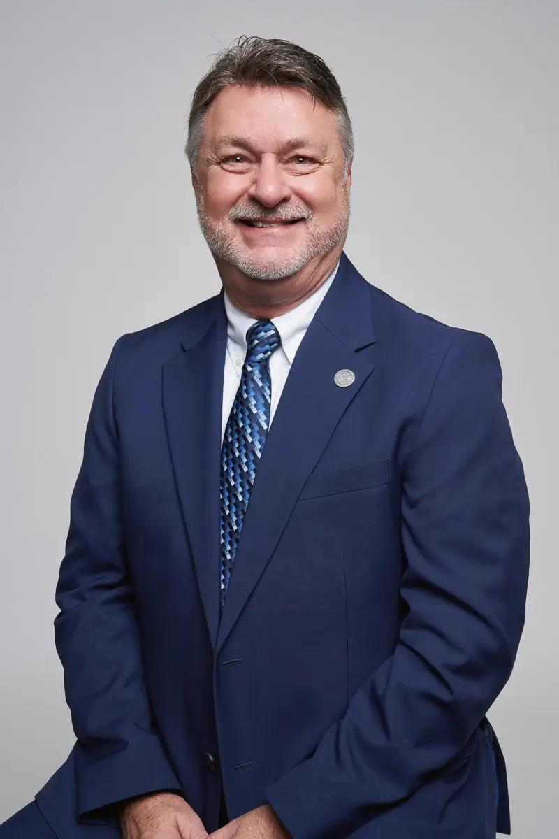 A middle-aged man wearing a blue suit, patterned tie, and white shirt poses for a professional portrait against a plain gray background. He is smiling and has short, gray-streaked hair and a trimmed beard.