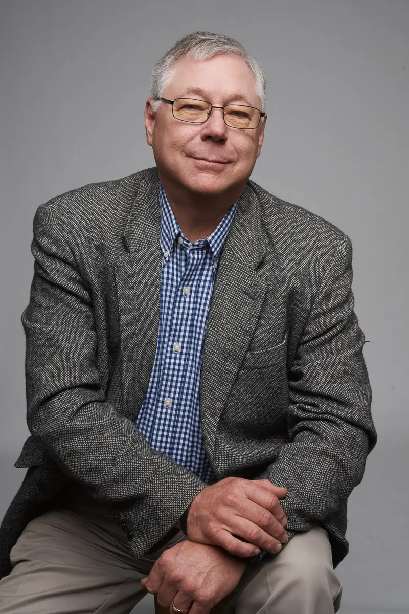 An older man with gray hair and glasses sits against a gray background, wearing a gray tweed blazer over a blue and white checkered shirt, with one hand resting on his knee and a slight smile on his face.