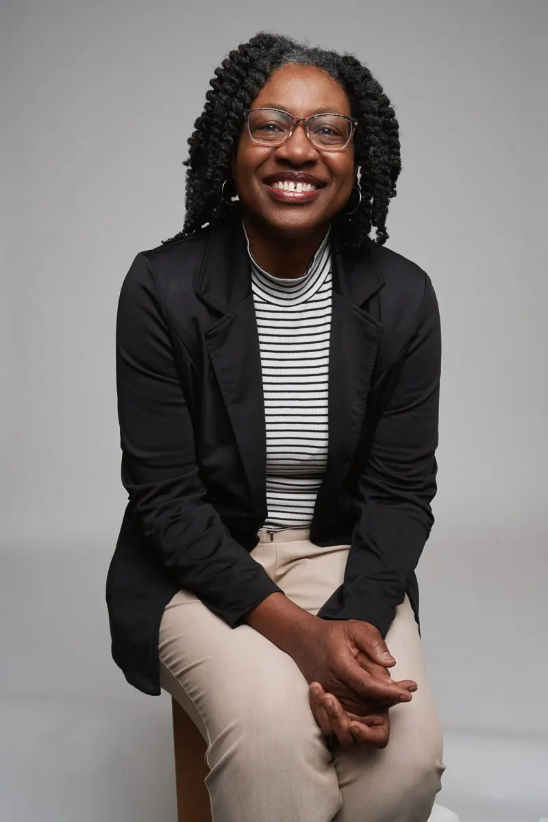 A person with glasses and natural curly hair sits on a stool, smiling. They are wearing a black blazer over a black-and-white striped shirt and light-colored pants, with hands resting in their lap against a plain background.