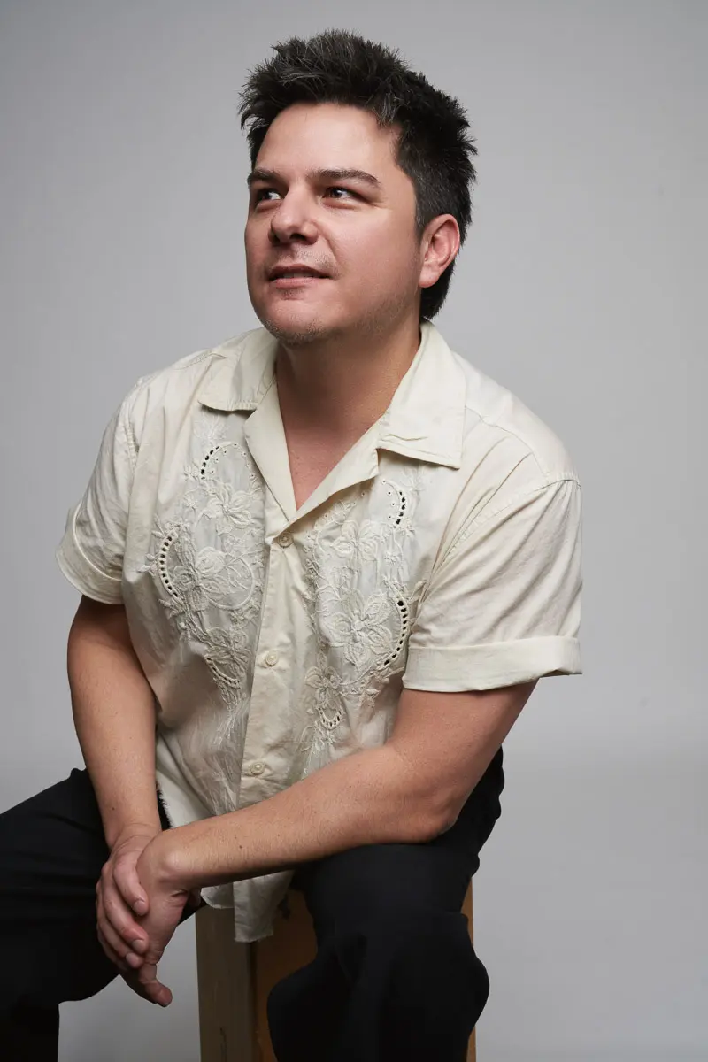 A person with short dark hair sits on a wooden block, wearing a cream-colored, short-sleeved shirt with embroidered floral patterns and black pants, looking slightly up and smiling against a plain gray background.