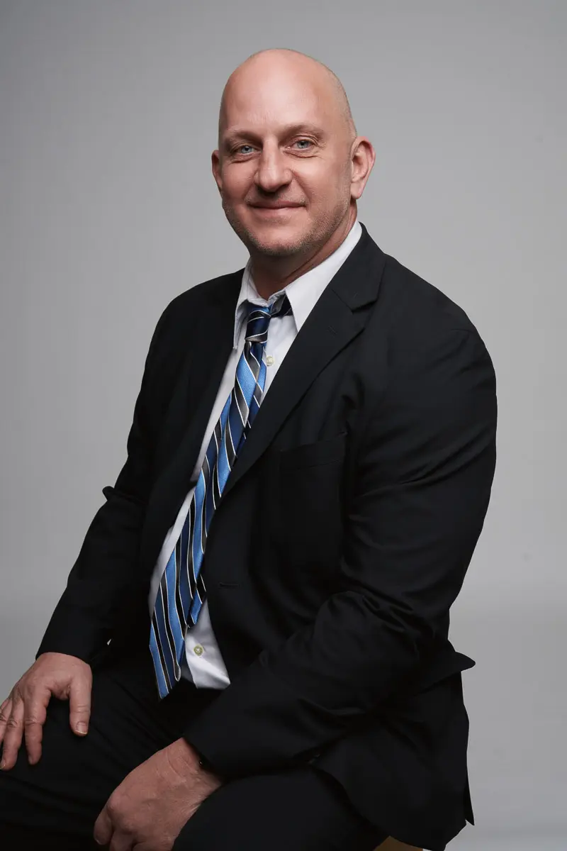 A bald man in a black suit, white shirt, and blue striped tie sits on a stool against a plain gray background, smiling at the camera.