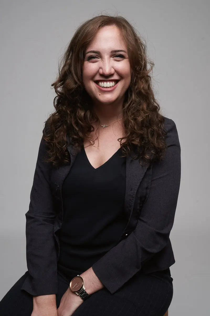A woman with curly brown hair, wearing a dark blazer and black top, sits on a stool against a plain light gray background, smiling warmly at the camera.