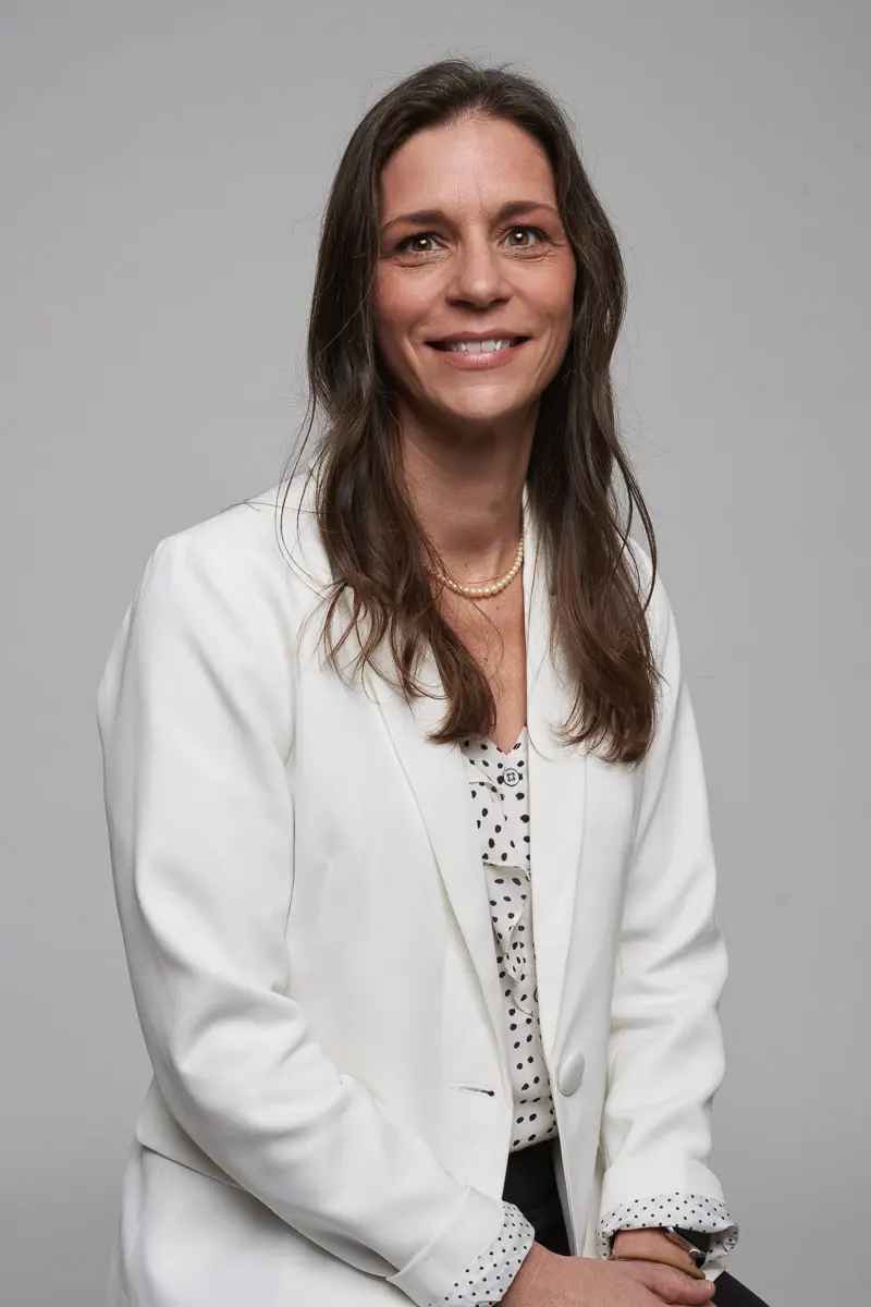 A woman with long brown hair, wearing a white blazer over a patterned blouse, sits and smiles at the camera against a plain gray background.