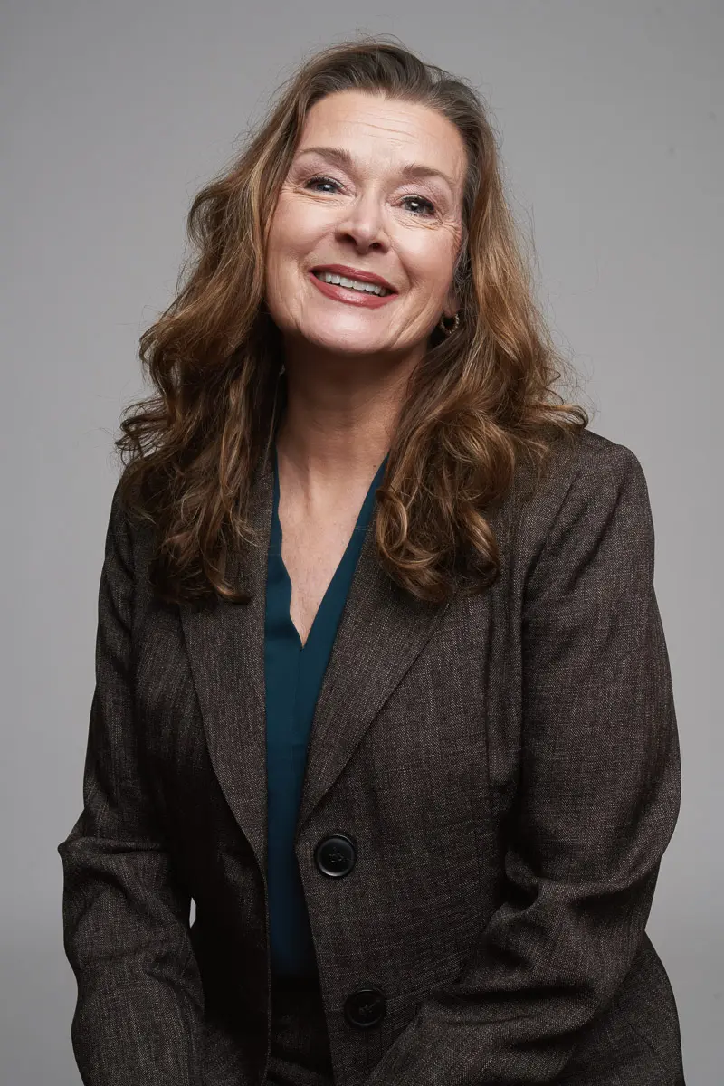 A woman with long wavy brown hair wearing a dark brown blazer and teal blouse smiles at the camera against a plain gray background.