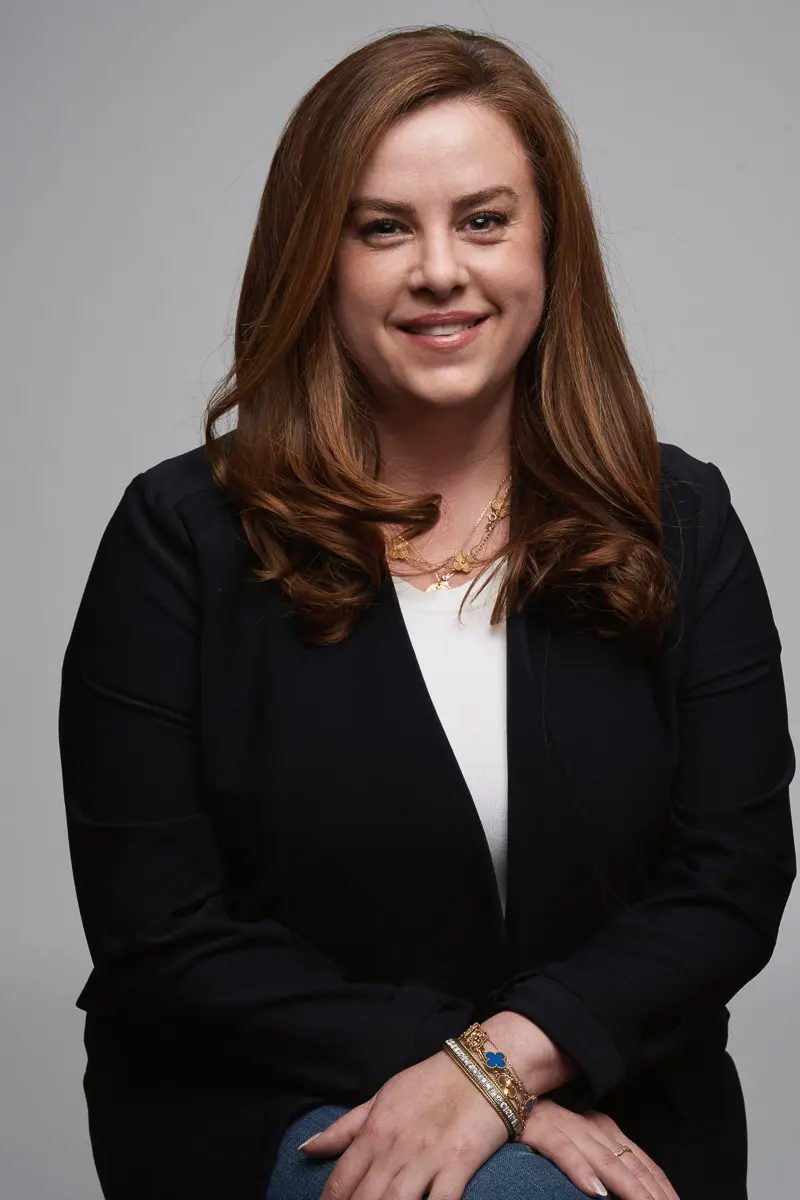 A woman with long brown hair wearing a black blazer, white top, and gold jewelry sits and smiles at the camera against a plain gray background.