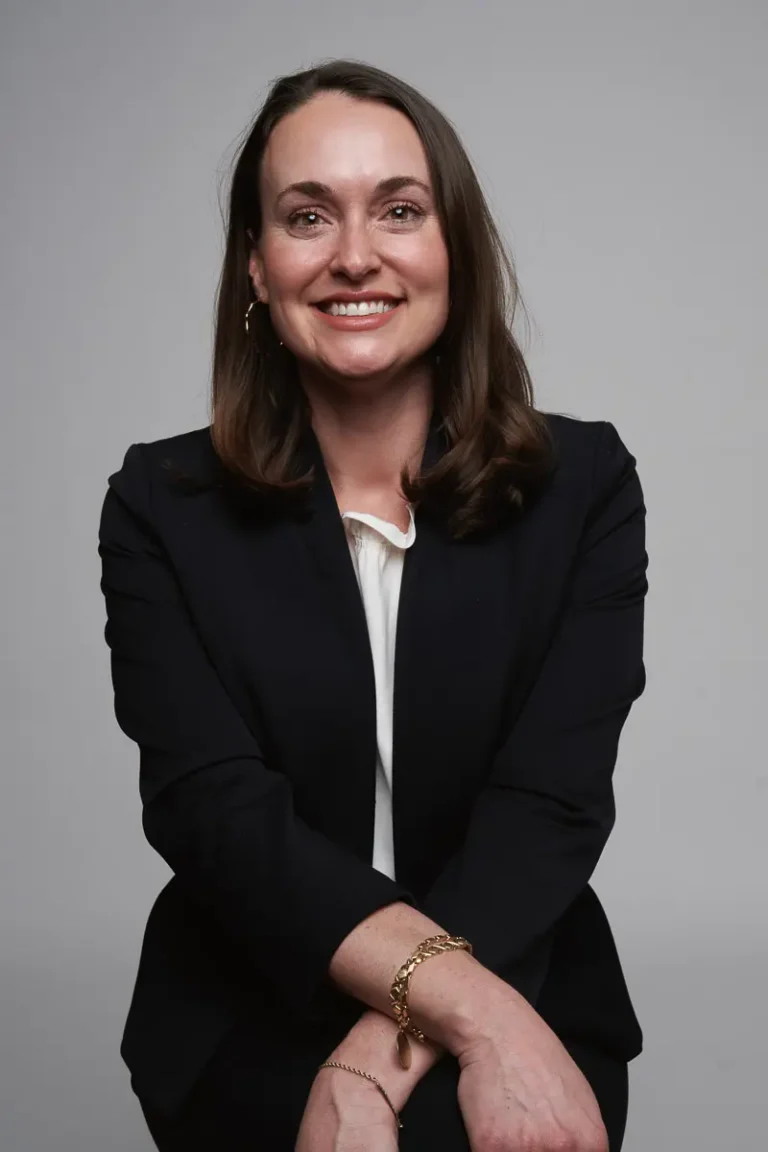 A woman with shoulder-length brown hair, wearing a dark blazer over a white blouse, sits and smiles at the camera against a plain gray background. Her hands are folded in her lap, and she wears a bracelet.