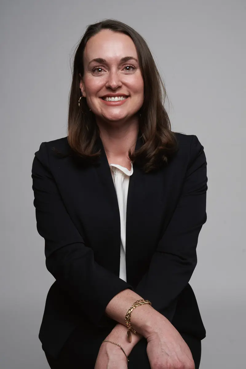 A woman with shoulder-length brown hair, wearing a dark blazer over a white blouse, sits and smiles at the camera against a plain gray background. Her hands are folded in her lap, and she wears a bracelet.