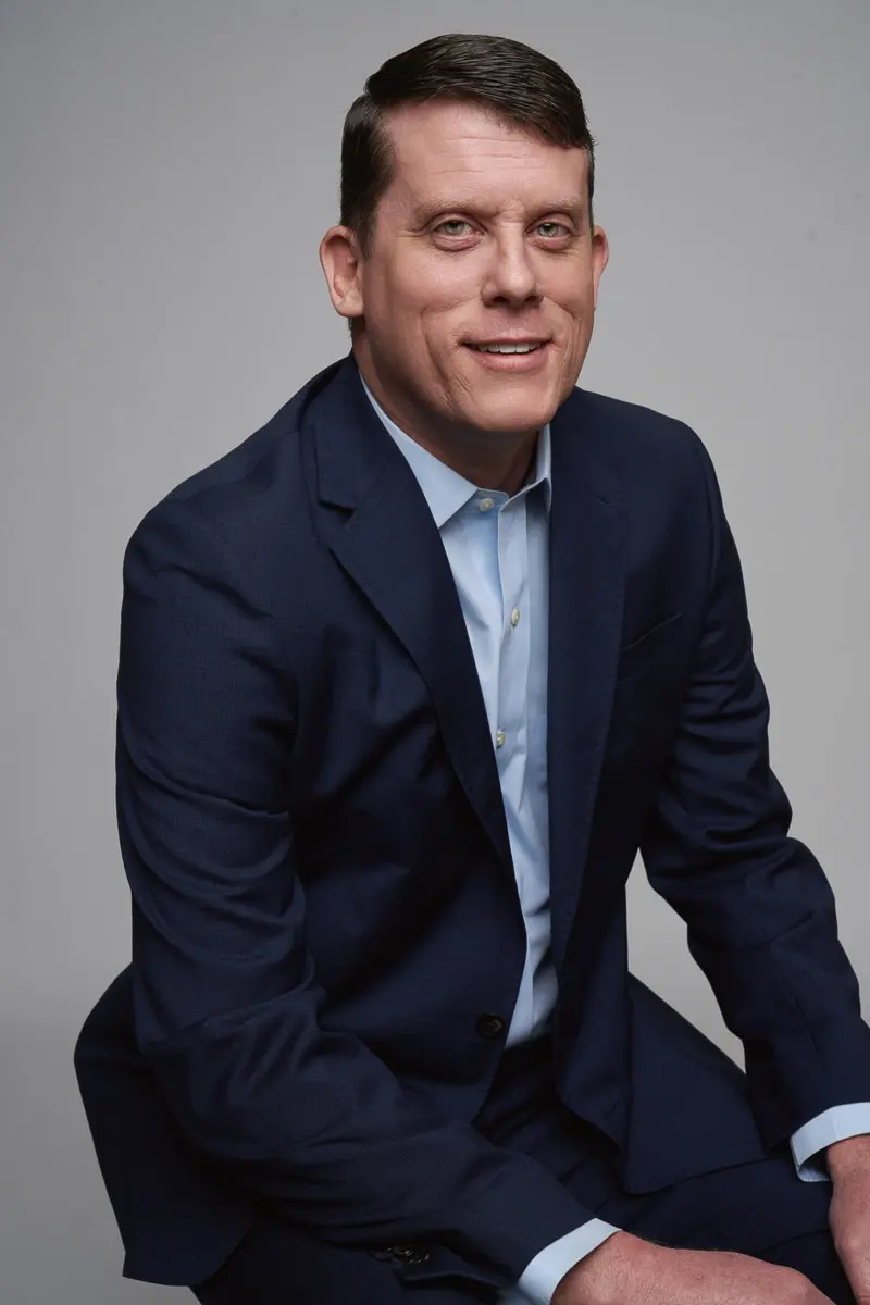 A man in a navy blue suit and light blue shirt sits and smiles at the camera against a plain, light gray background.