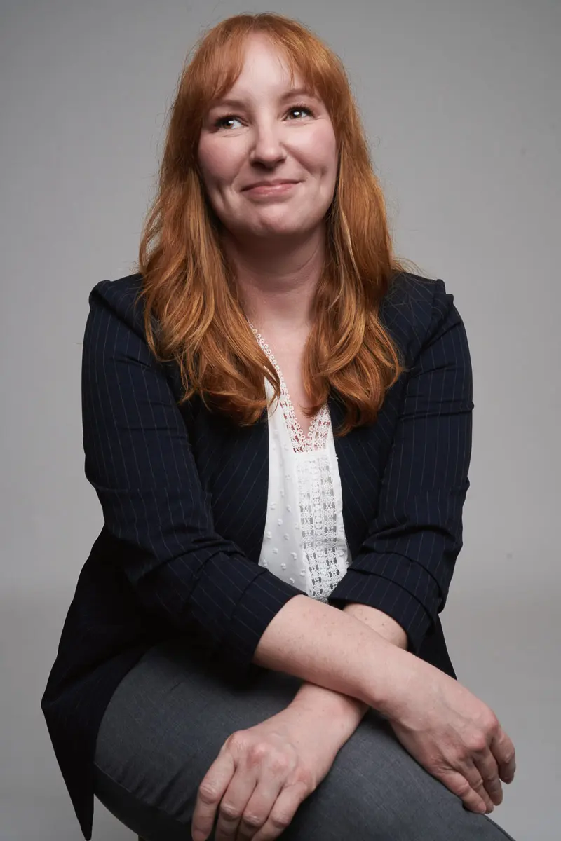 A woman with long red hair wearing a navy blazer and white blouse sits with her arms crossed on her knee, smiling softly and looking slightly up, against a plain gray background.