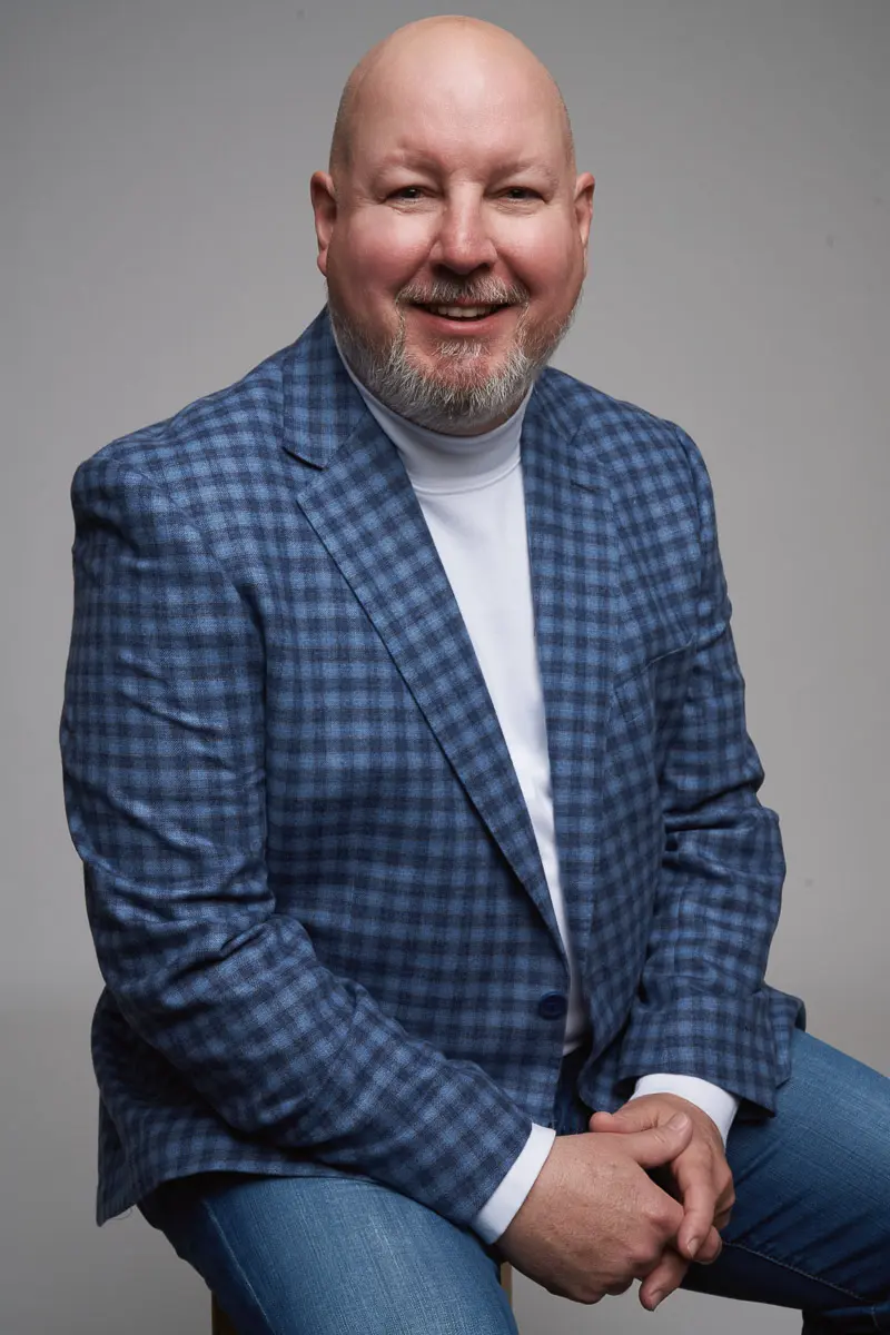 A smiling bald man with a beard wears a blue plaid blazer, white turtleneck, and jeans, sitting on a stool against a plain light gray background.
