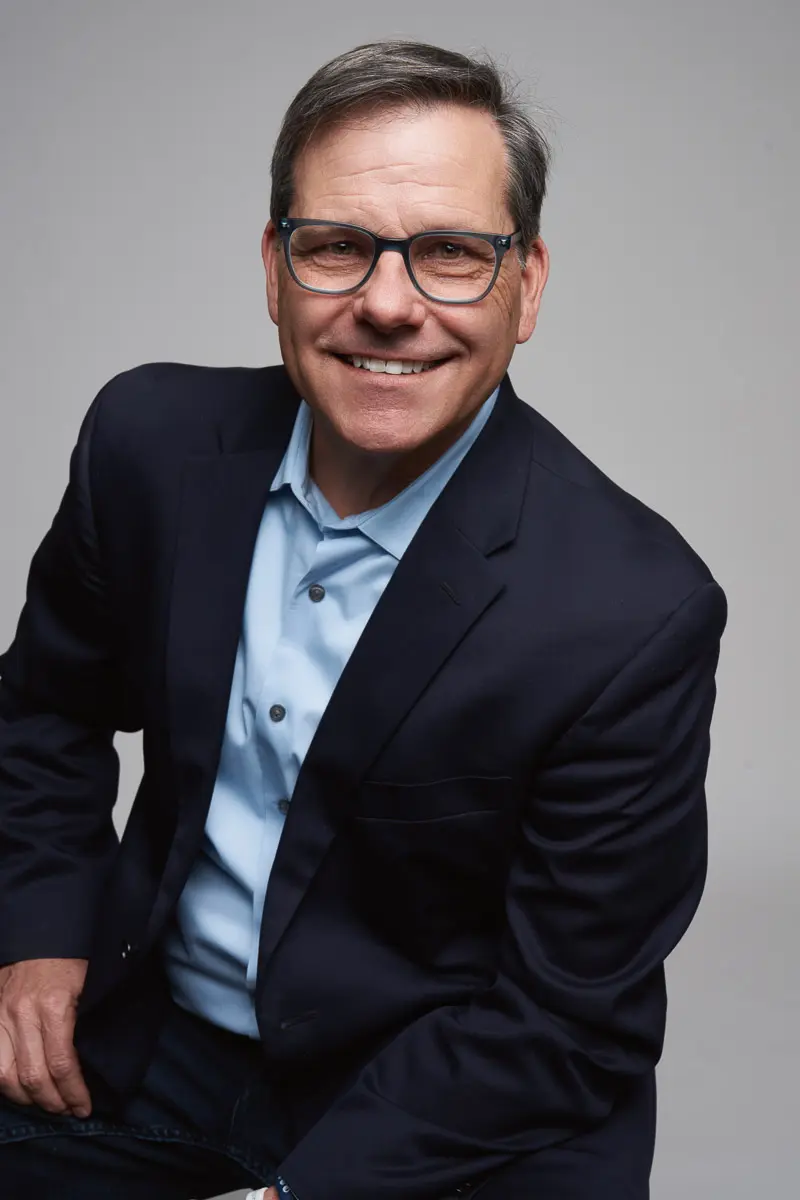 A middle-aged man with short brown hair and glasses smiles at the camera. He is wearing a dark blazer over a light blue shirt and is seated against a plain gray background.