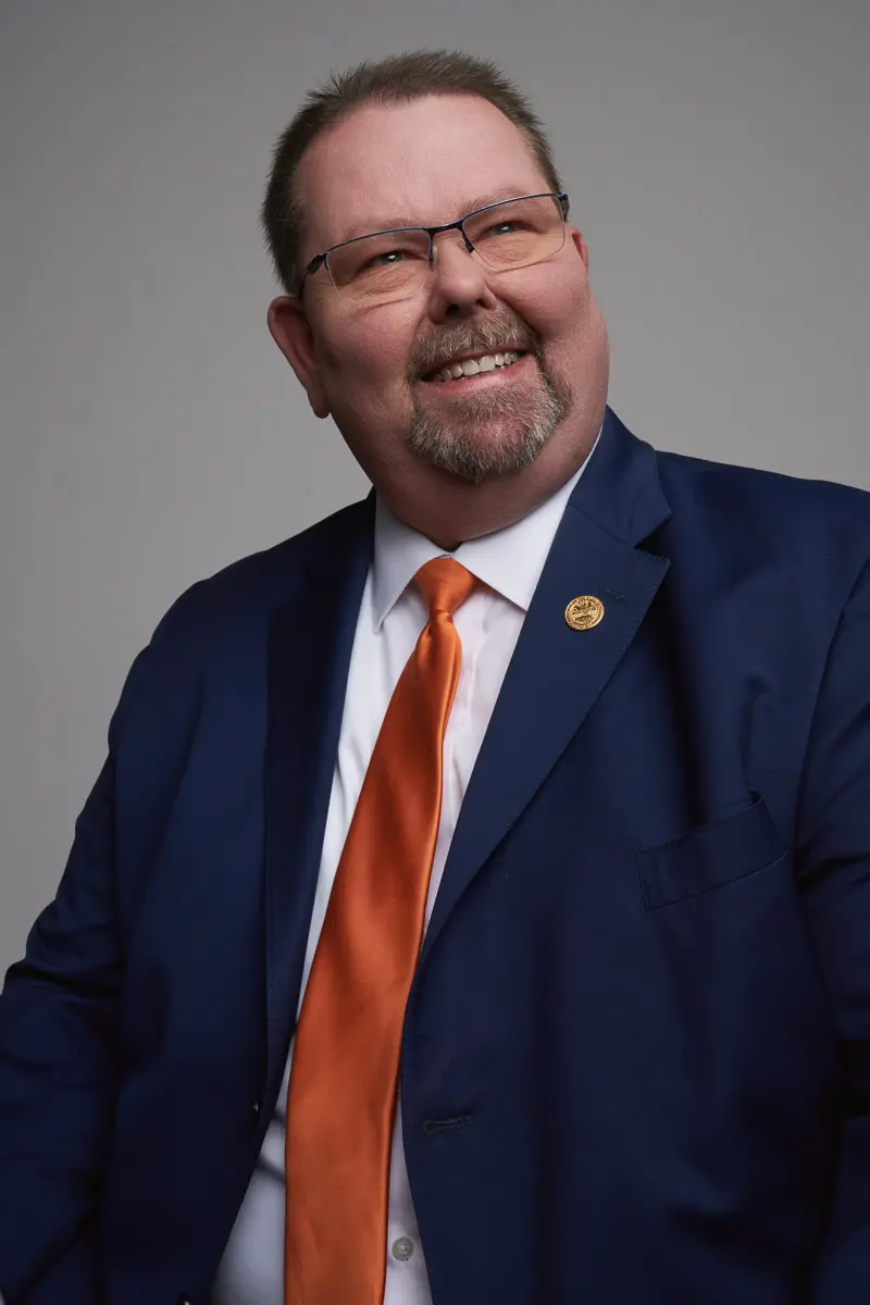 A man wearing glasses, a dark blue suit, a white shirt, and an orange tie smiles while looking slightly to the side. He has short hair, a beard, and a pin on his lapel, with a neutral gray background.