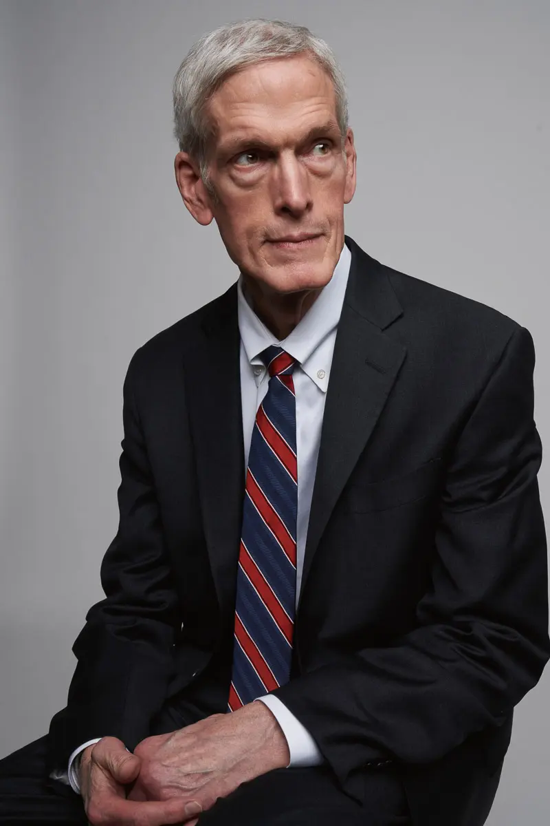 A mature man with short gray hair wearing a black suit, white dress shirt, and a red, white, and blue striped tie sits against a plain gray background, looking thoughtfully to his left.