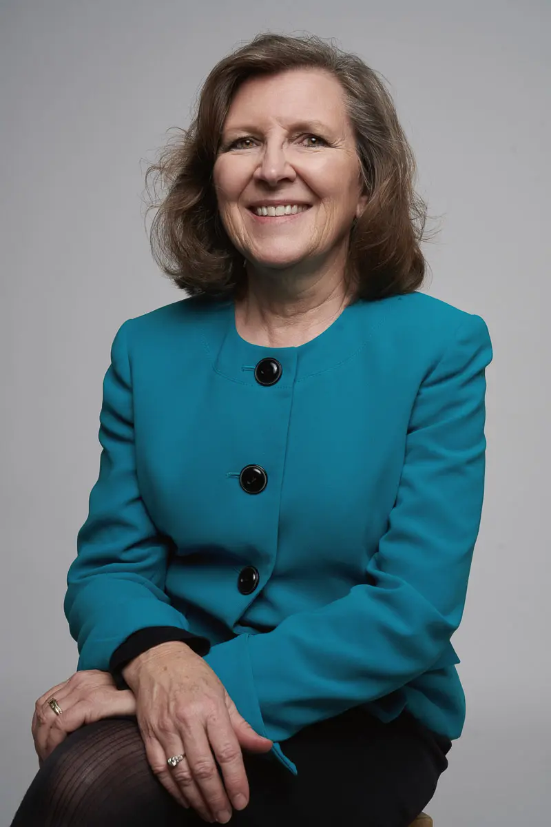 A woman with shoulder-length brown hair smiles while sitting, wearing a teal blazer with black buttons and black pants, against a plain light gray background.