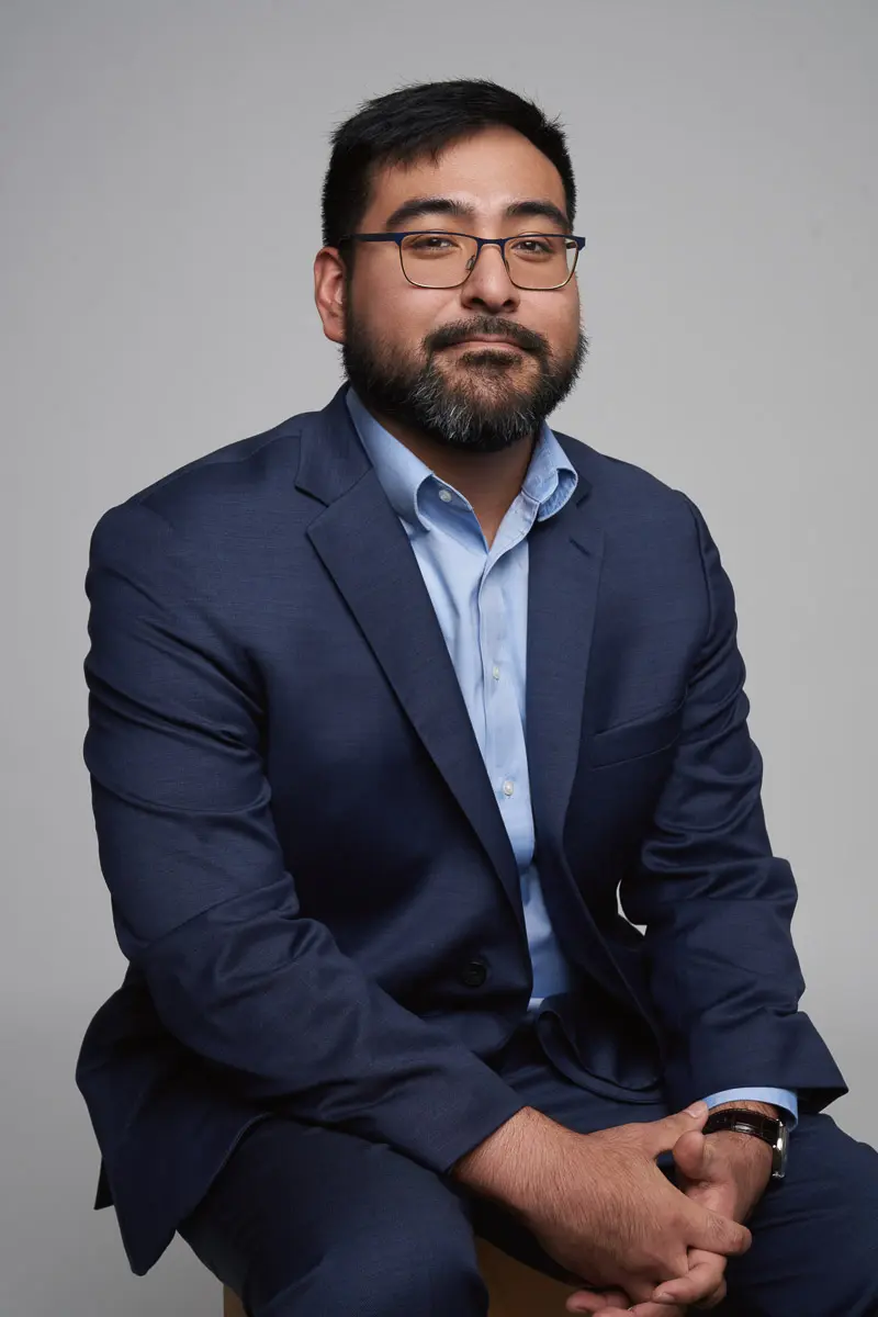 A man with glasses and a beard, wearing a navy blue suit and light blue shirt, sits on a stool with his hands resting on his knees, looking at the camera against a plain gray background.