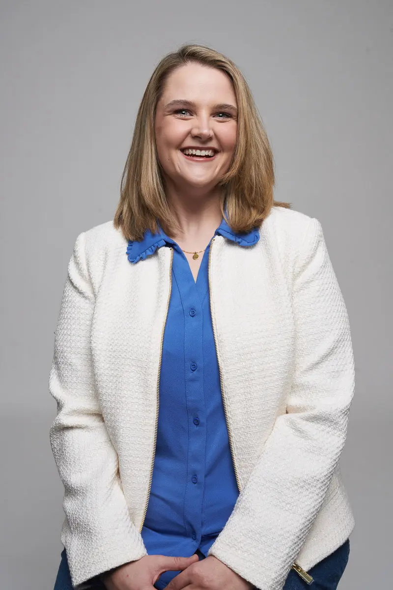 A woman with shoulder-length blonde hair is smiling and seated against a plain gray background. She is wearing a white textured jacket over a blue blouse and has her hands resting in her lap.