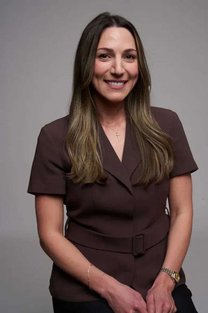 A woman with long brown hair wearing a brown short-sleeve blazer, gold watch, and bracelet, smiles at the camera against a plain gray background.