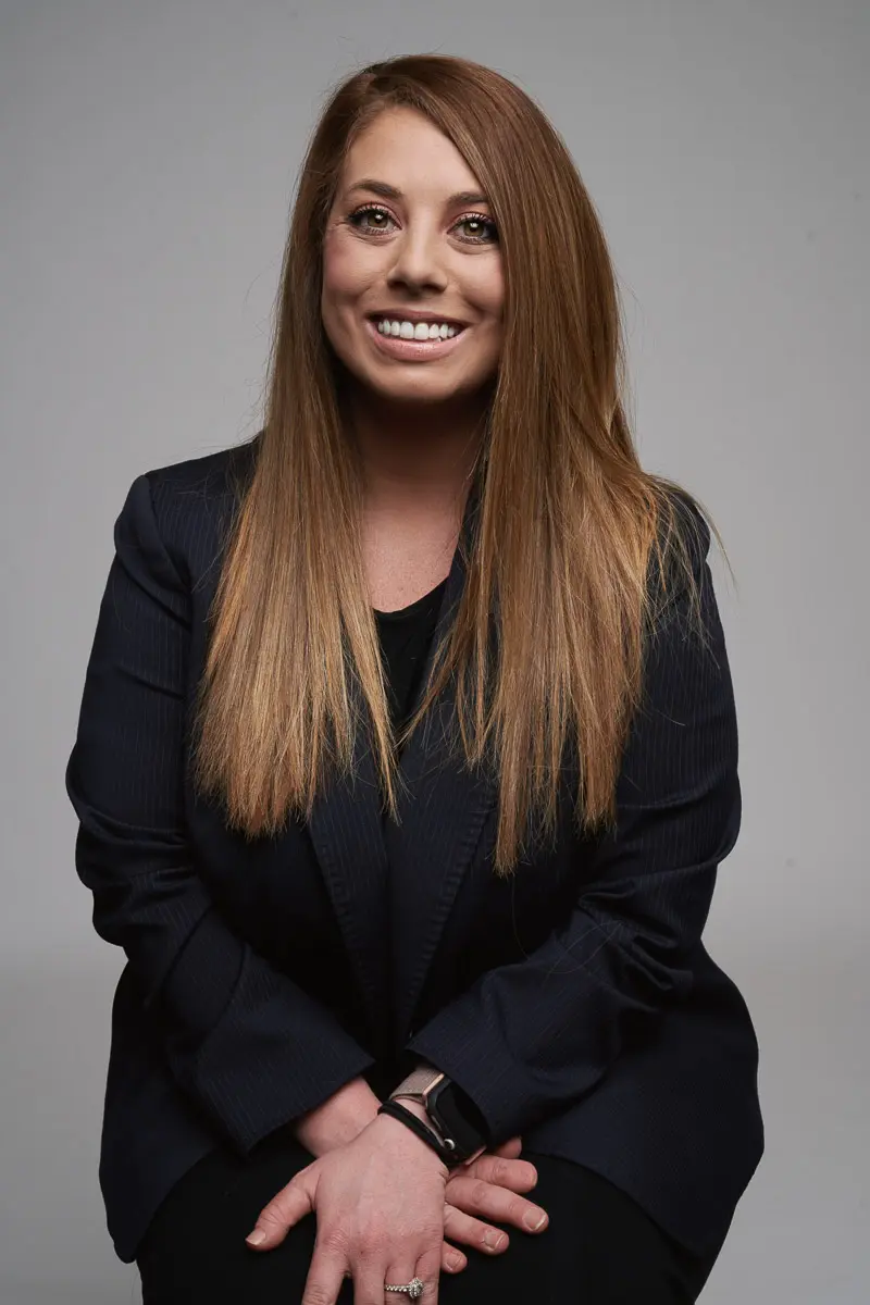 A woman with long straight light brown hair, wearing a dark blazer over a black top, smiles while sitting with hands folded in her lap against a plain gray background.