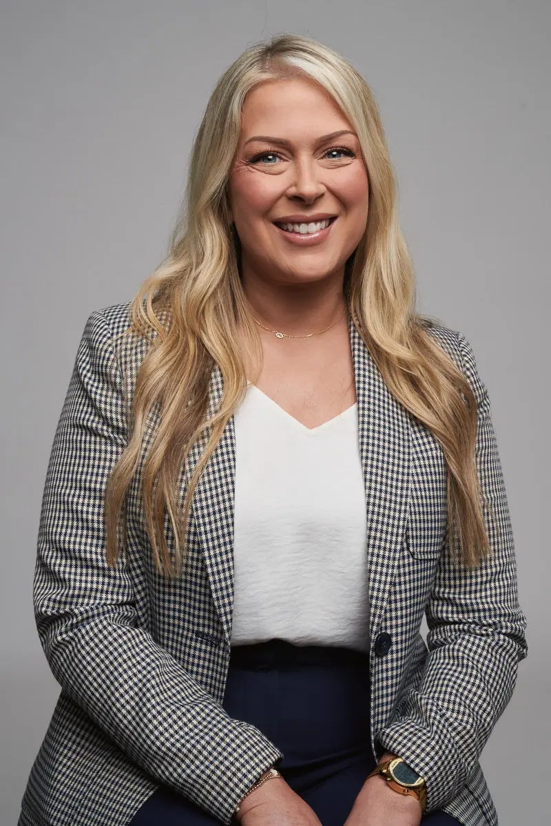 A woman with long blonde hair, wearing a checked blazer, white blouse, and dark pants, sits and smiles at the camera against a plain gray background.