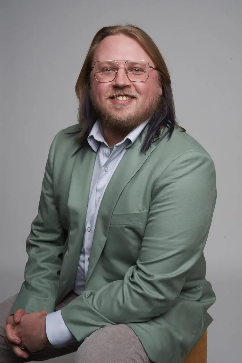 A person with long light brown hair, glasses, and a beard is sitting on a stool, smiling. They are wearing a light green blazer over a light blue shirt and light-colored pants, with a neutral gray background.