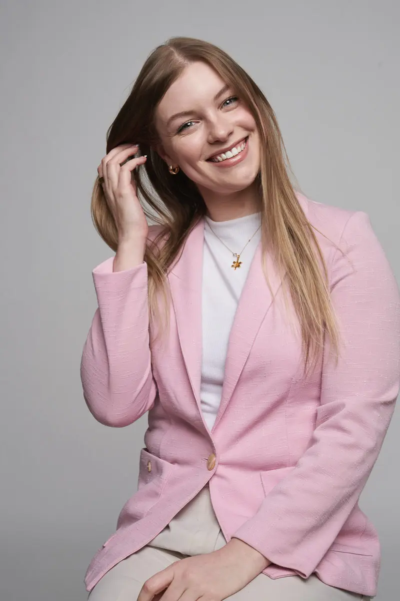 A smiling woman with long blonde hair wears a light pink blazer over a white top. She is seated, touching her hair with one hand, and looks directly at the camera against a plain gray background.