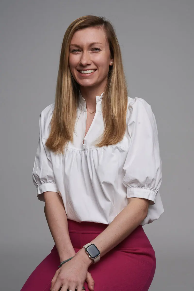A woman with long blonde hair, wearing a white blouse and pink pants, sits and smiles at the camera. She has a smartwatch on her wrist and poses against a plain gray background.
