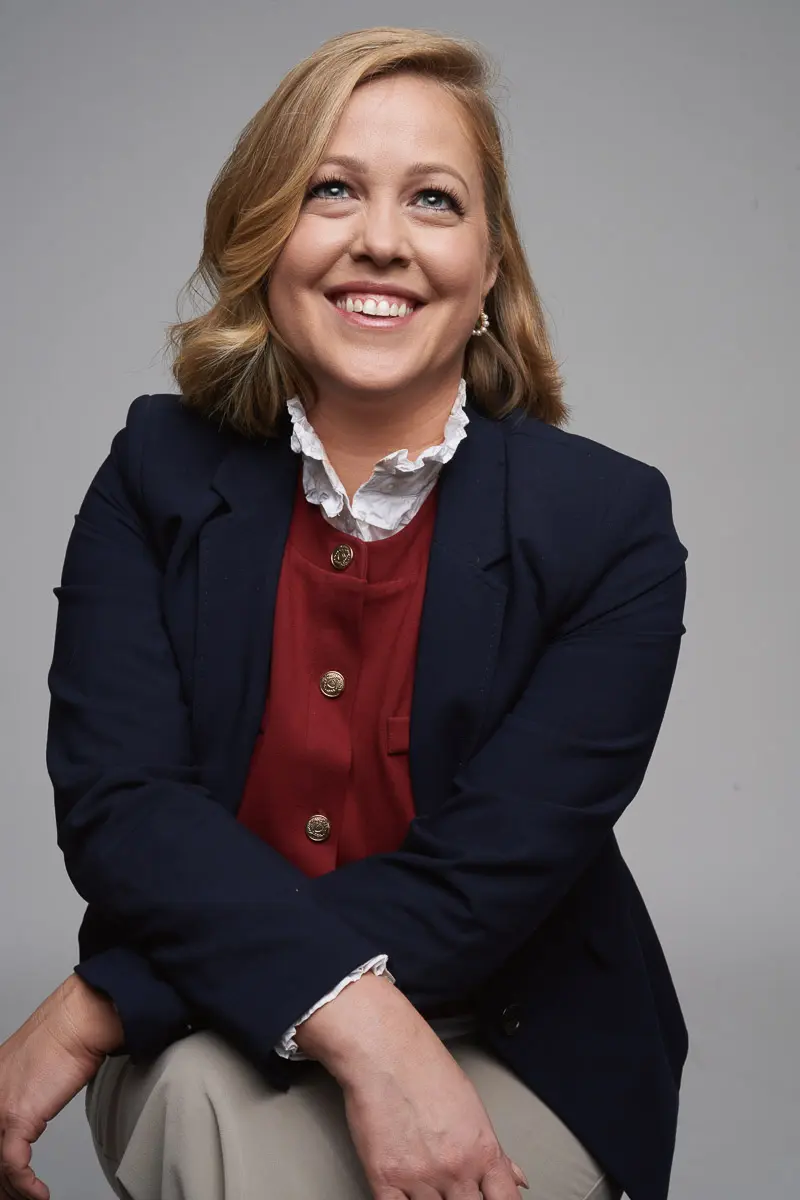 A smiling woman with blonde hair sits against a plain background, wearing a navy blazer over a red blouse with ruffled collar and beige pants. She looks upward with a cheerful expression.
