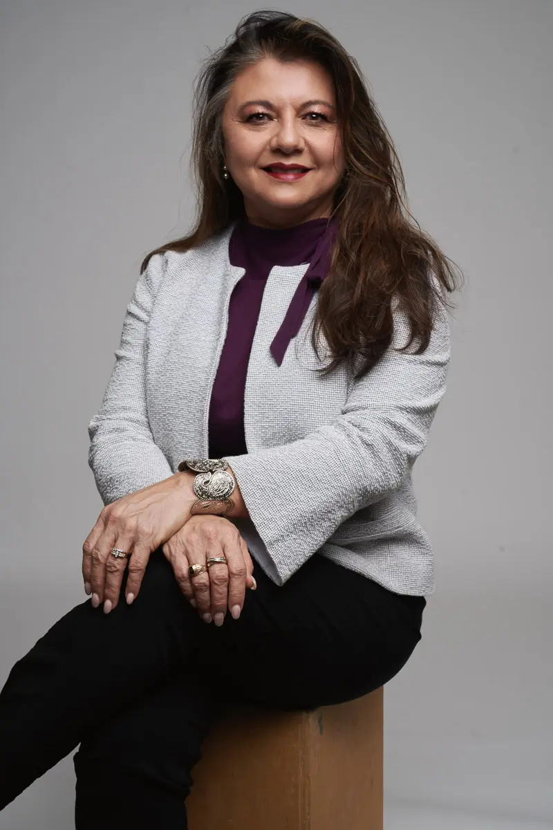 A woman with long brown hair sits on a wooden stool, smiling. She wears a light gray jacket over a purple top, black pants, and jewelry including rings and a watch. The background is plain light gray.