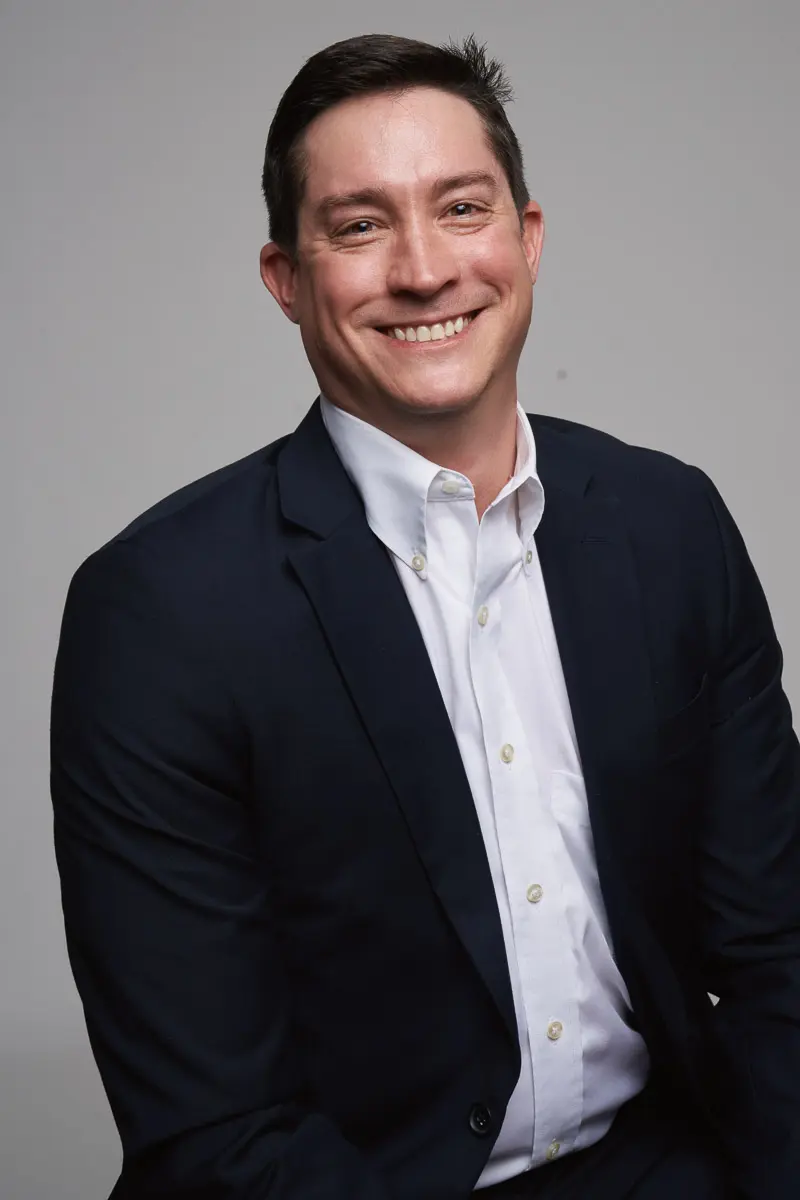 A man with short dark hair smiles while wearing a dark suit jacket over a white dress shirt, sitting against a plain, light gray background.