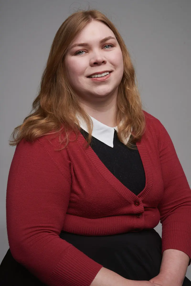 A person with long, light brown hair wearing a red cardigan over a black top with a white collar, sitting and smiling in front of a gray background.