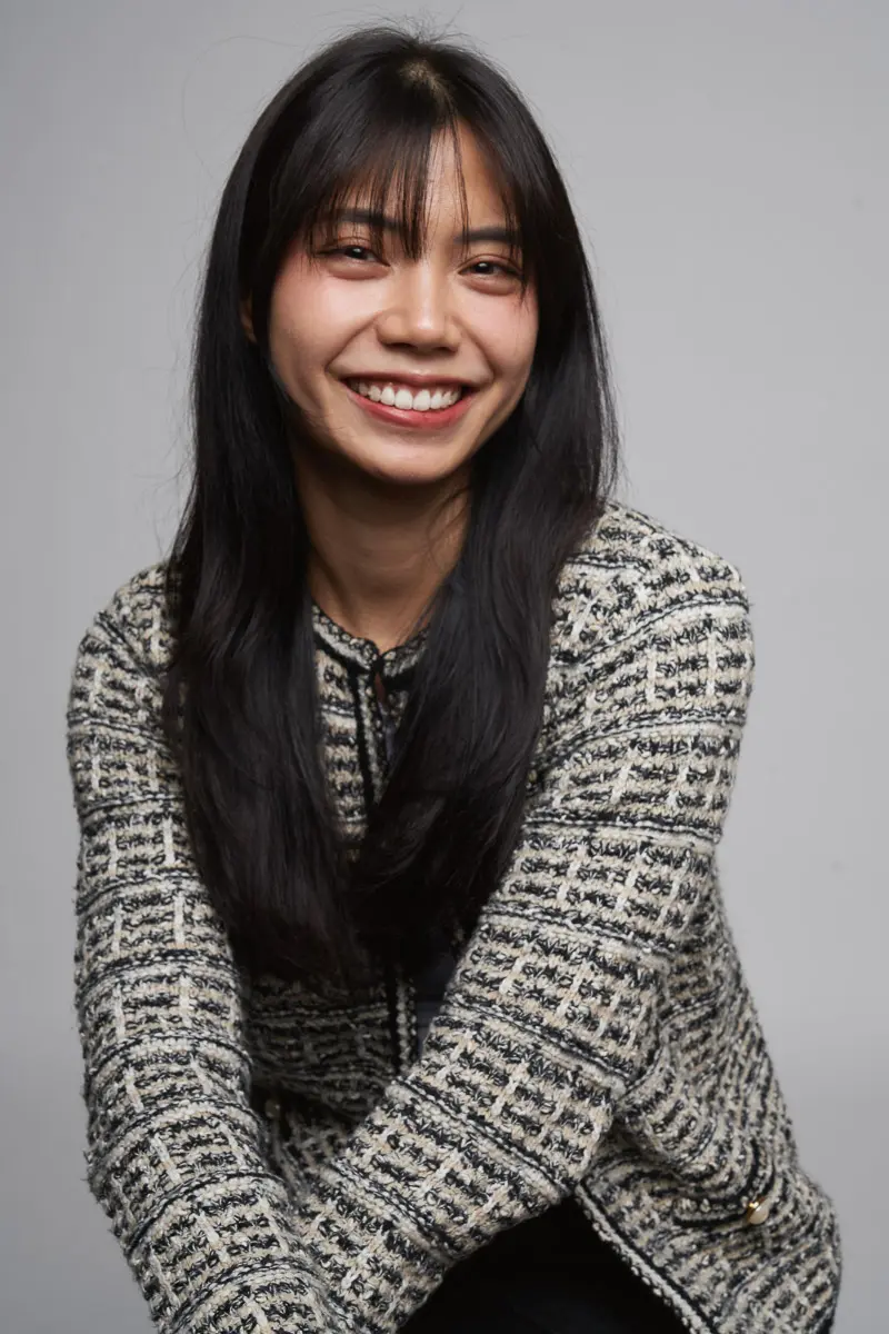 A young woman with long, straight black hair smiles at the camera. She is wearing a textured, patterned jacket and sits against a plain, light gray background.