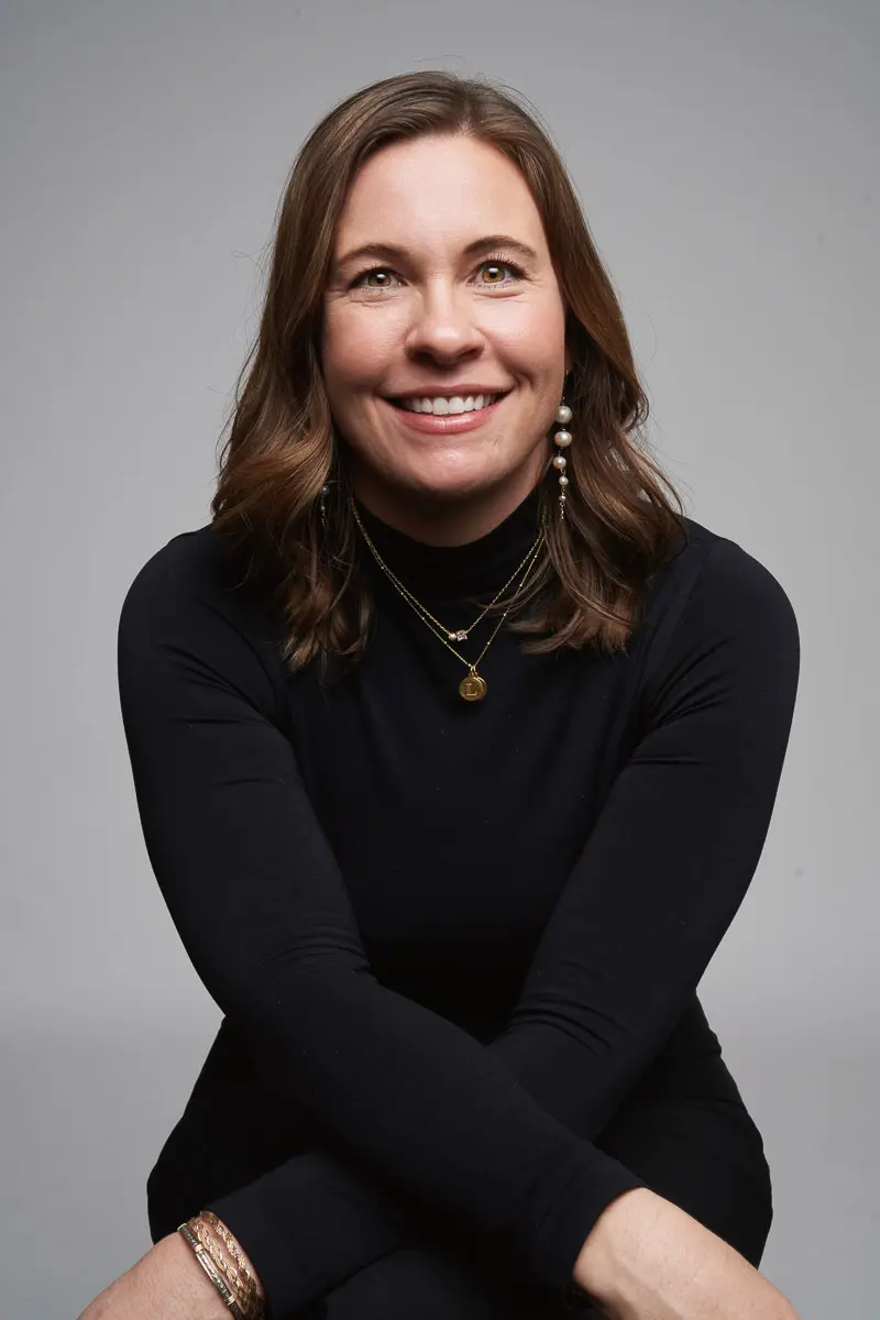 A woman with shoulder-length brown hair, wearing a black long-sleeve top, layered necklaces, and earrings, sits with her arms crossed and smiles at the camera against a plain gray background.