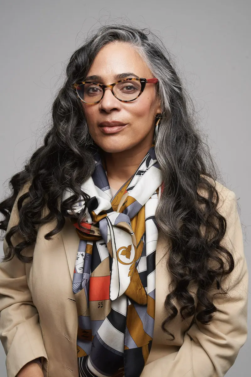 A woman with long, wavy gray hair wears glasses, a tan blazer, and a colorful patterned scarf. She faces forward with a neutral expression against a plain gray background.
