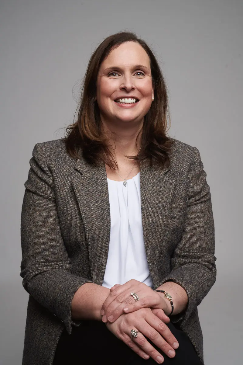A woman with shoulder-length brown hair is smiling while sitting with her hands clasped. She is wearing a grey blazer over a white blouse and has rings on her fingers. The background is plain and light grey.