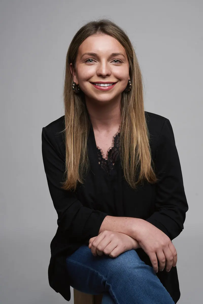 A woman with long, straight blonde hair sits on a stool against a plain gray background. She wears a black blouse with lace trim and blue jeans, smiling warmly with her hands on her knee—an elegant portrait in the style of Scottie Tudor.