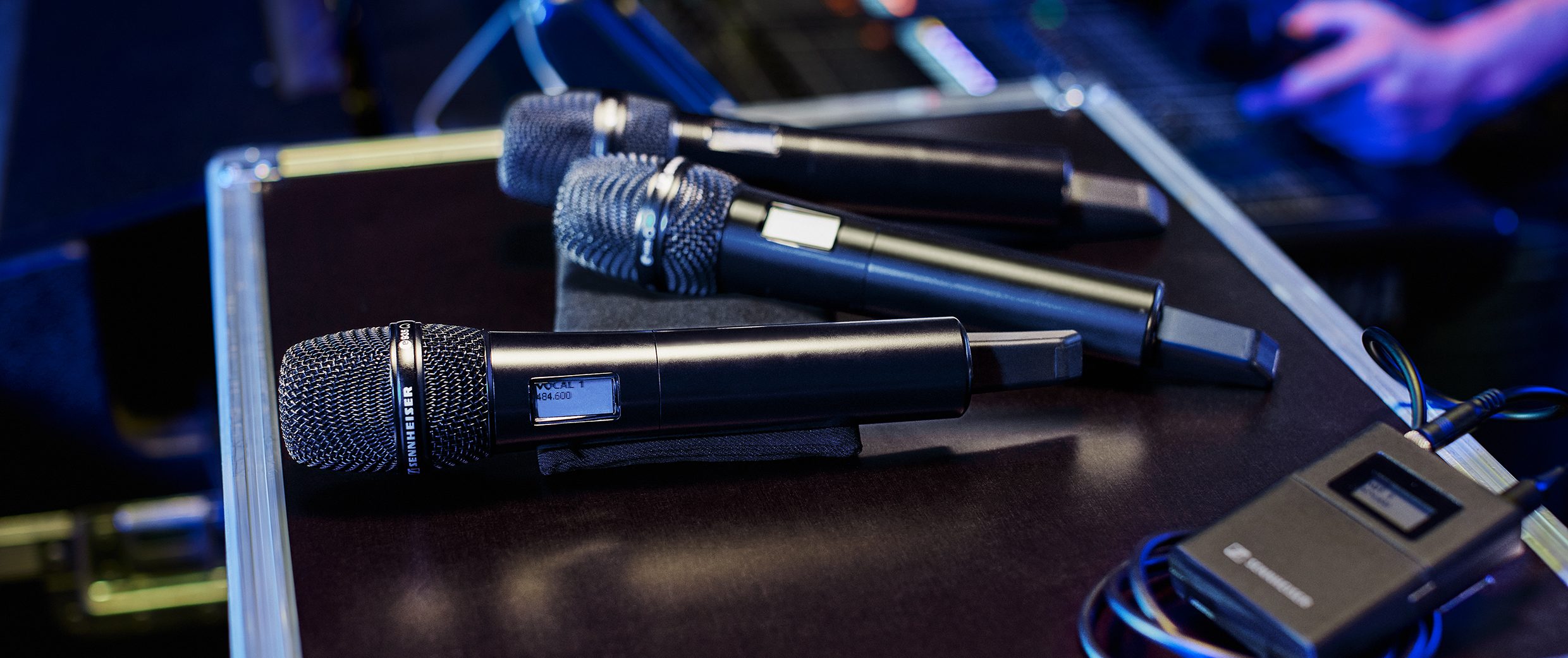 Three wireless microphones are lying on a black surface next to a wireless transmitter, with blurred equipment and a person’s hand visible in the background. The scene suggests a stage or studio setting.