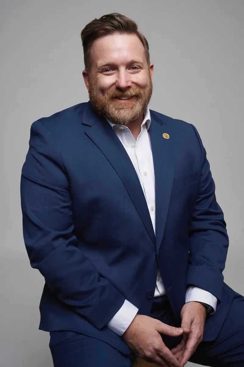 A man with light skin, a beard, and short hair is wearing a blue suit and white shirt. He is sitting and smiling at the camera against a plain, light gray background.