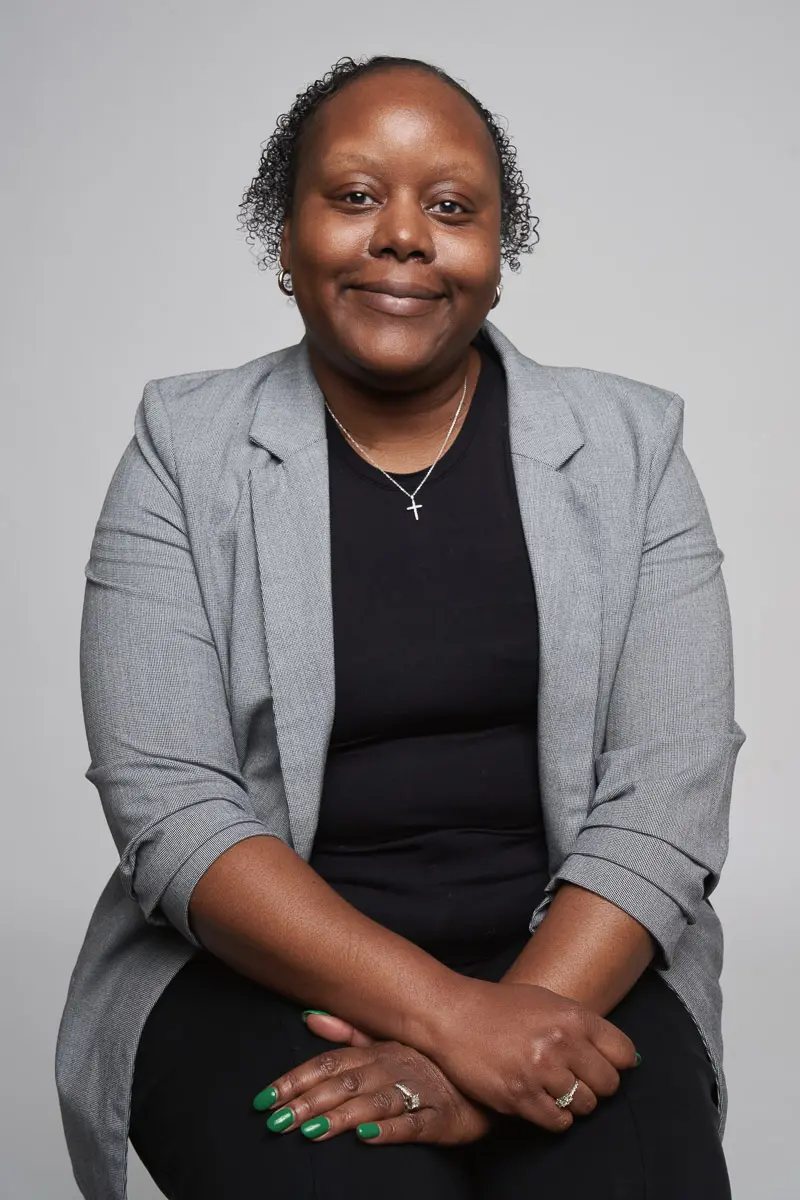 A woman with short curly hair, wearing a gray blazer over a black top, sits with her hands folded on her lap. She is smiling softly and has green nail polish, a ring, and a cross necklace. The background is plain and light gray.