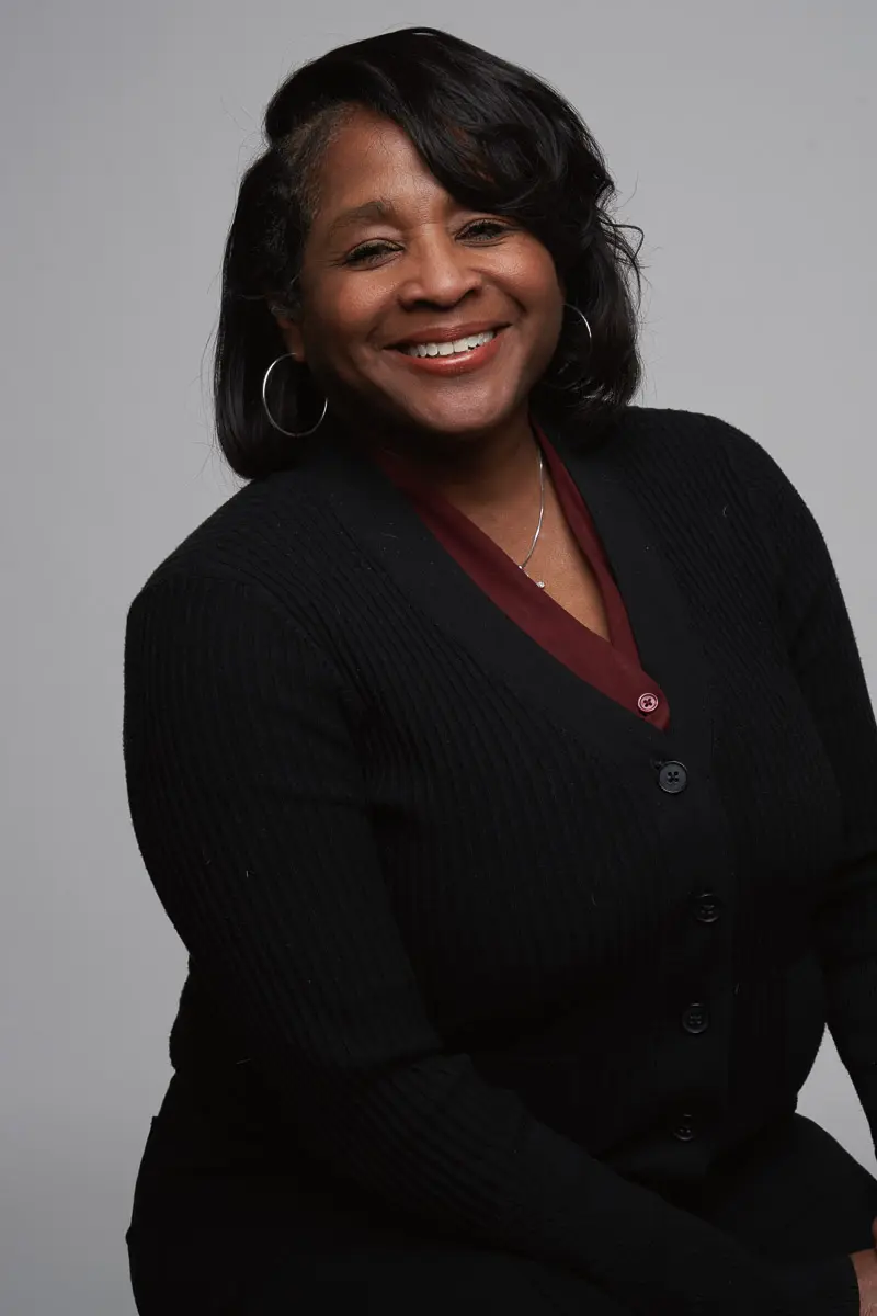 A woman with shoulder-length dark hair is smiling, wearing a black cardigan over a burgundy top with hoop earrings, sitting against a plain light gray background.