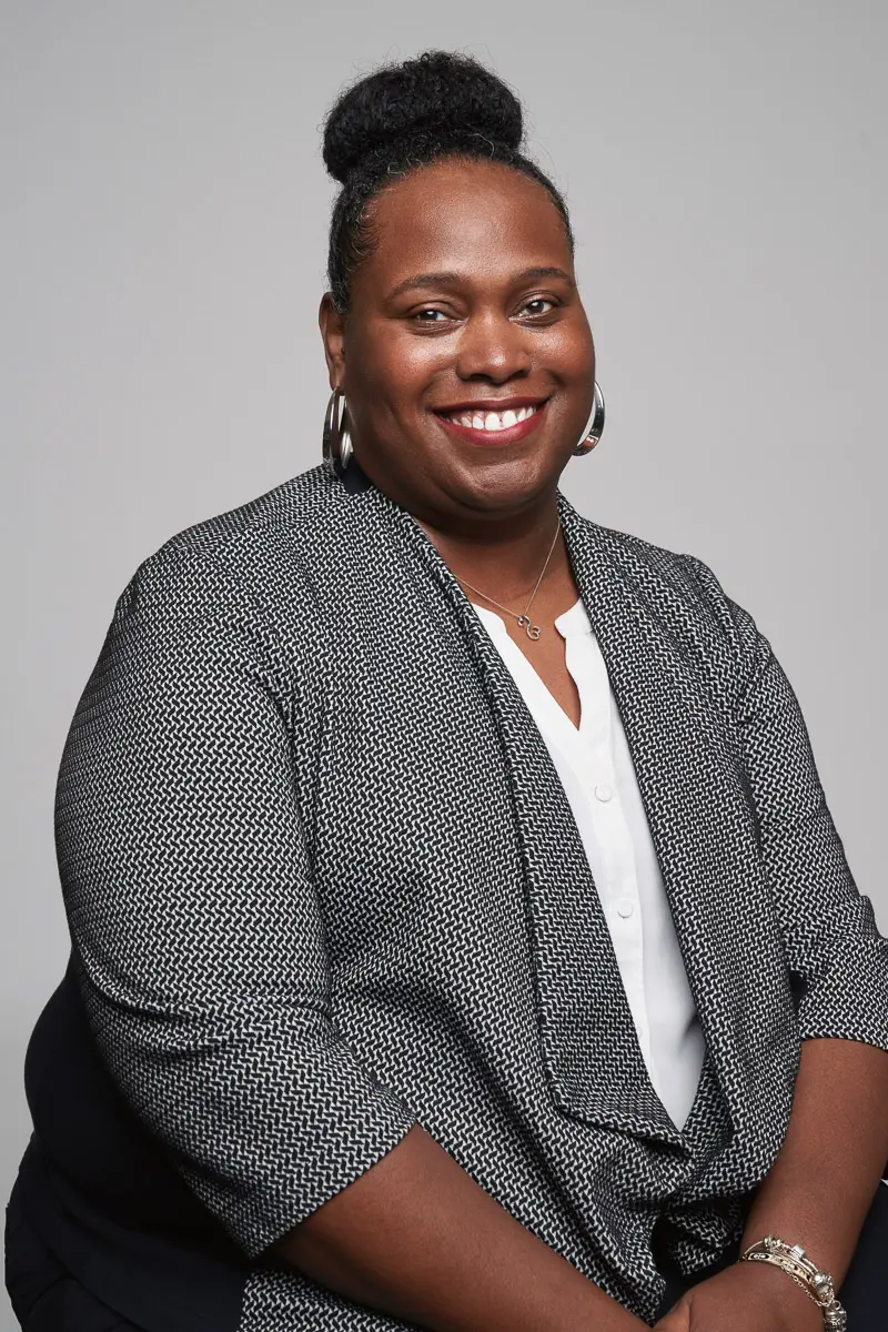 A smiling woman with dark hair in a bun, wearing a patterned blazer over a white blouse, silver hoop earrings, and a bracelet, sits against a plain light gray background.